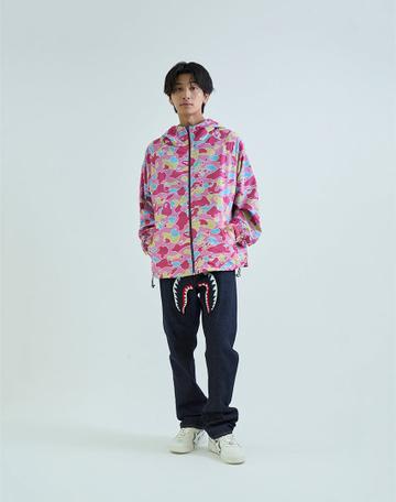 A man wearing a zipped pink multicolor camo hooded jacket, rigid dark indigo jeans with a red-and-white shark mouth embroidered across the upper thighs, and white leather sneakers, standing against a pale grey background.