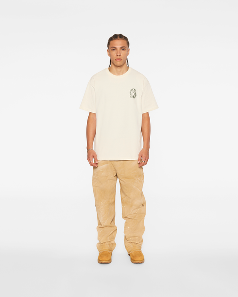 A man wearing an oversized cream Billionaire Boys Club t-shirt with a small astronaut-helmet logo on the chest, baggy paint-flecked tan canvas carpenter pants stacked over wheat nubuck work boots, standing head to toe on a white background.