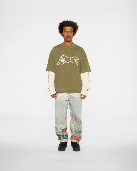 A man wearing an oversized olive Ice Cream T-shirt with a white running-dog graphic layered over a cream long-sleeve top with bone-print sleeves, loose light-wash jeans printed with a vintage ice-cream-parlour street scene, and black lace-up boots, standing against a white background.