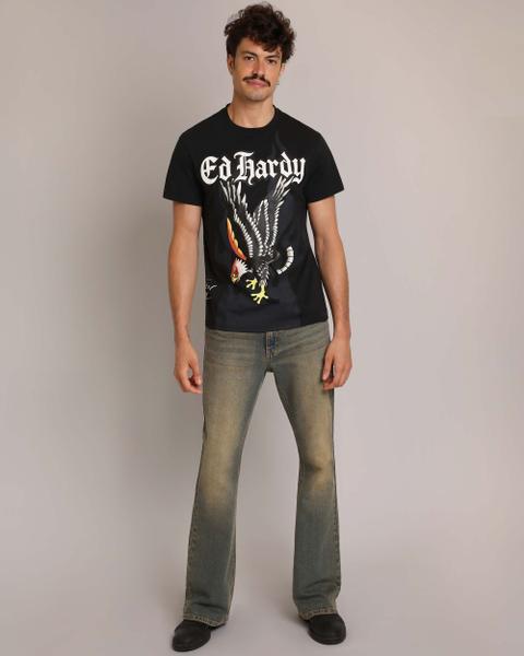 A man wearing a black Ed Hardy t-shirt printed with a tattoo-style eagle and gothic script, faded tinted bootcut jeans and black boots, standing on a grey studio background.