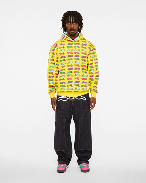 A man wearing a yellow full-zip hoodie, zipped up and all-over printed with multicoloured running dogs, with wide raw-indigo double-knee jeans featuring a white running-dog graphic at the waistband, and pink-and-green suede sneakers, standing head to toe on a white background.
