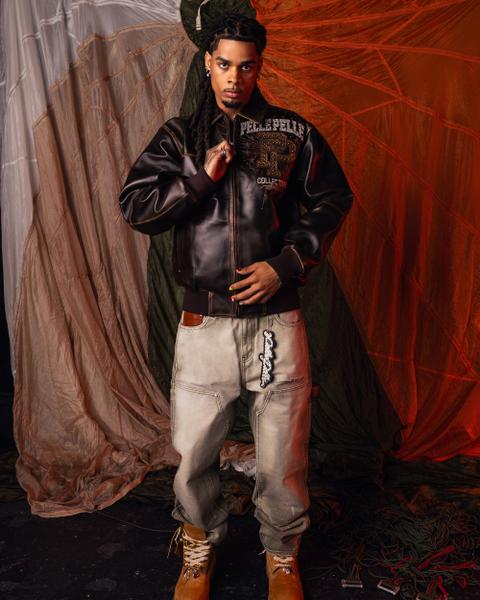 A man wearing a dark brown leather Pelle Pelle bomber with a studded PP crest, baggy washed grey carpenter jeans stacked over wheat nubuck work boots, and a rhinestone script keychain at the hip, standing against draped parachute silk.