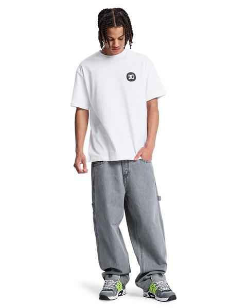 A man wearing an oversized white t-shirt with a small black DC logo on the chest, very baggy light grey carpenter jeans with a hammer loop whose hems pile over chunky grey, black and neon-green padded skate shoes, standing head to toe on a white background.